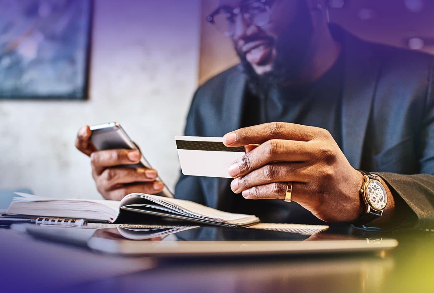 Man with glasses looks at cell phone while holding a credit card in the other hand.