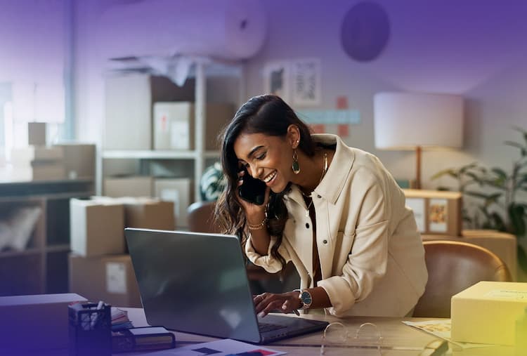 Woman on phone while typing on laptop, leaning over desk with lots of boxes and shipping materials behind her.