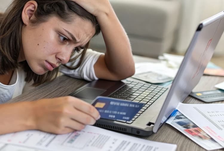 Frustrated woman looking at laptop while holding a credit card and surrounded by bills.